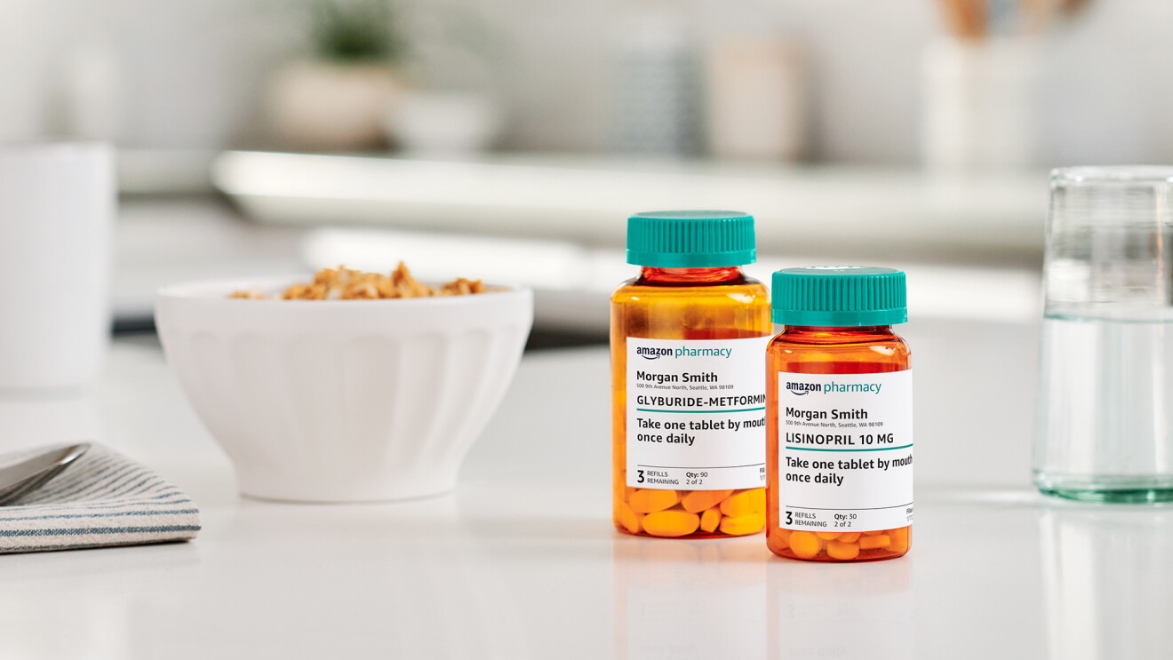 Two Amazon Pharmacy pill bottles sit on a white kitchen countertop in between a glass of water and a bowl of cereal.