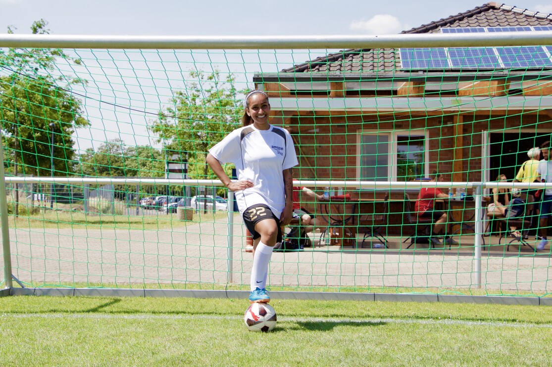 Eine dunkelhäutige Fußballerin vor einem Tor: Sie trägt ein weißes Sportshirt, ihr Fuß ist locker auf einem Ball abgelegt.