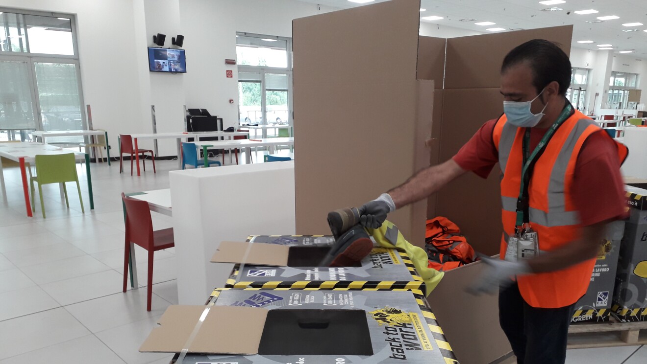 Scatole per il riciclo di indumenti all'interno di un'area comune di un magazzino Amazon. A destra, un operatore di magazzino, con indosso la mascherina e il giubbino catarinfrangente, sta introducendo in una delle due scatole delle scarpe da lavoro.