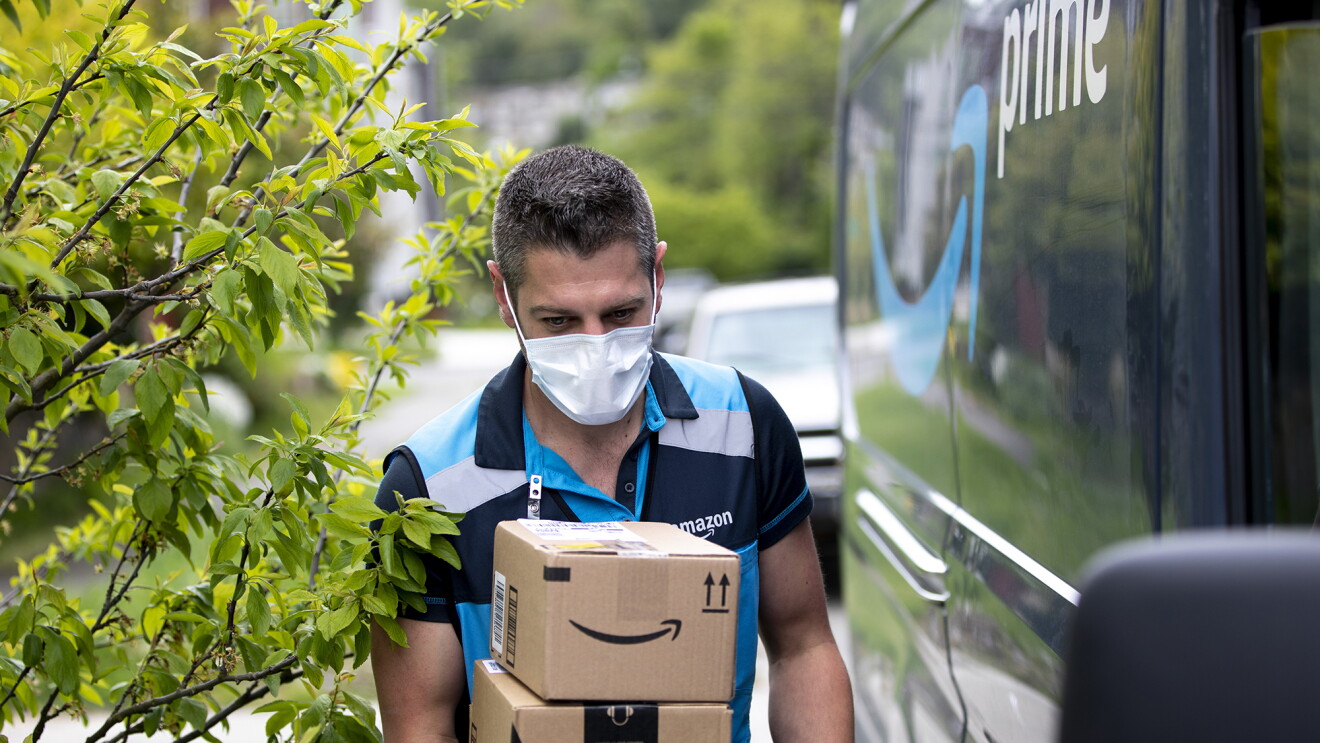 A delivery driver wearing a face mask walks alongside an Amazon Prime delivery van, carrying packages.