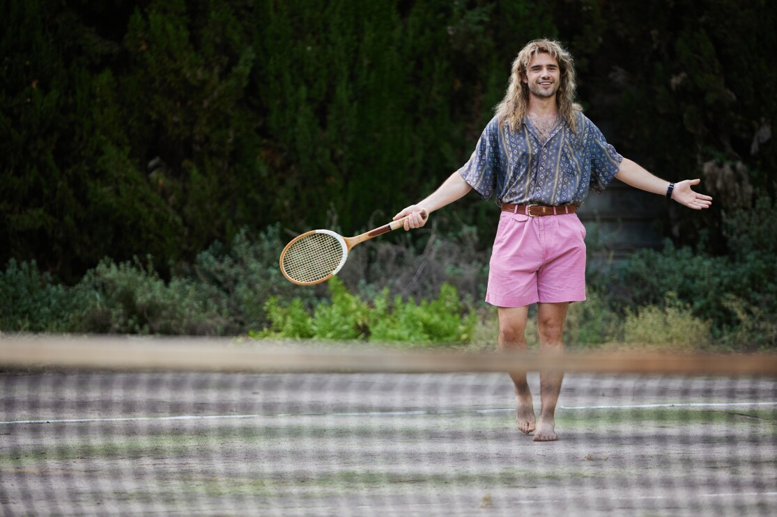 Szene aus Perfect Match, der Geschichte über Steffi Graf und Andre Agassi. Schauspieler Toby Sebastian trägt in seiner Rolle als Andre Agassi pink-farbene Shorts zu einem gemusterten Hemd. Er ist barfuß auf einem Tennis-Court, hält in der einen Hand einen Tennisschläger.