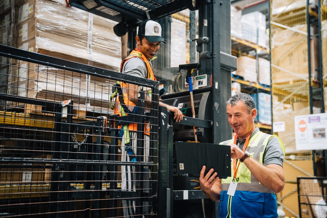 Temporary associate John Sheerin smiling at a colleague in an Amazon Fulfilment Centre and typing on a tablet.