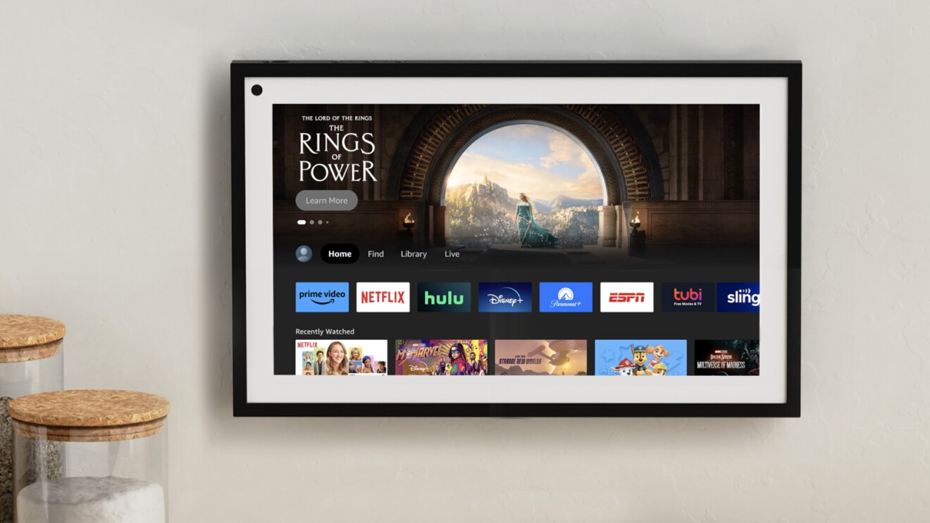 An image of an Amazon Echo Show 15 mounted on the wall showing the homepage with a "The Lord of The Rings Rings of Power" promo at the top and miscellaneous apps at the bottom.