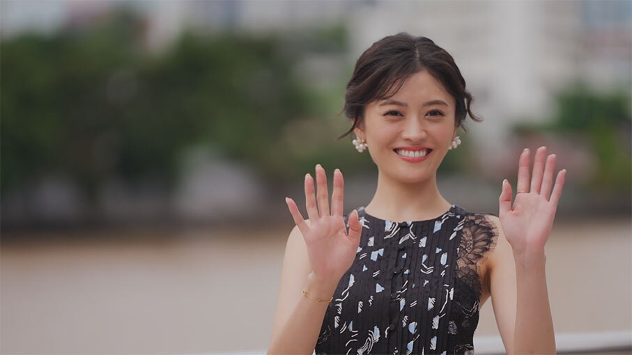 背後に建物や木々が見える屋外で、笑顔の女性が肩のあたりまで上げた両手を振っている。