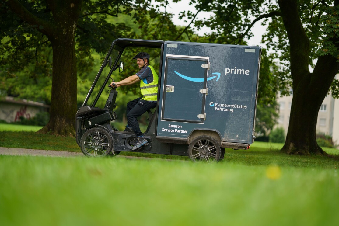 Ein junger Fahrer in einem mit Amazon gebrandeten E-Cargo Bike: Er trägt Berufskleidung im Amazon-Look und ist mit einem blau-grauen E-Lastenrad unterwegs.