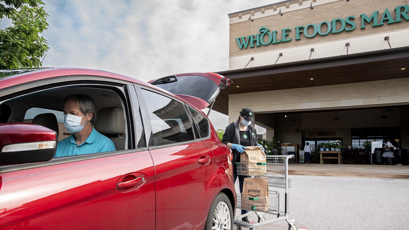 一个男人戴着口罩接收在线杂货从整个食品市场秩序,送到他的汽车后备箱里。