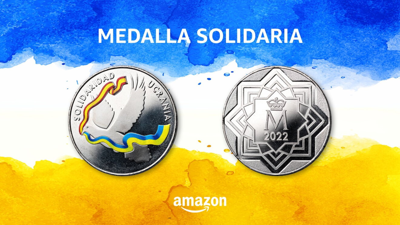 Medalla solidaria Ucrania. Sobre un fondo con los colores de la bandera de Ucrania, de color azul y amarillo, pero con una franja blanca, aparece las dos caras de la medalla. En la cara principal el escrito: Solidaridad Ucrania y una silueta de paloma que se su pico salen en color las banderas de España y Ucrania. El reverso de la medalla tiene el año 2022 y el logo de la Fábrica Nacional de Moneda y Timbre. En la parte superior con letras blancas y mayúsculas Medalla Solidaria y abajo Amazon y la sonrisa. 
