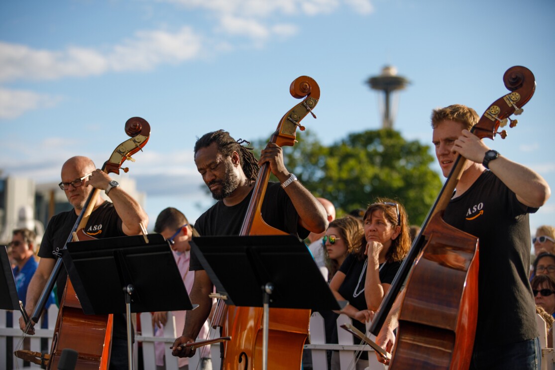 Three members of the Amazon Symphony Orchestra are seen playing their cellos. Each wears a black tee shirt, and stands behind a music stand. Behind the musicians, a crowd of attendees is visible.