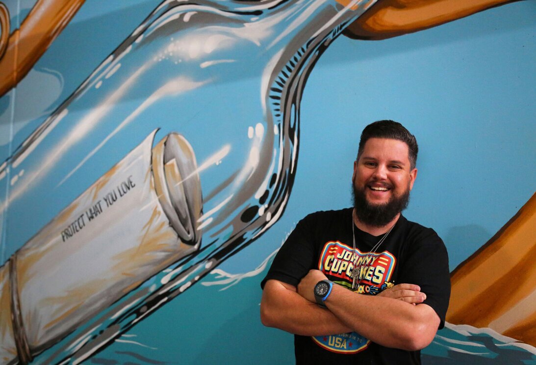 An image of Patrick Maxcy smiling for a photo in front of a mural he painted of a bottle with a folded up paper in it.