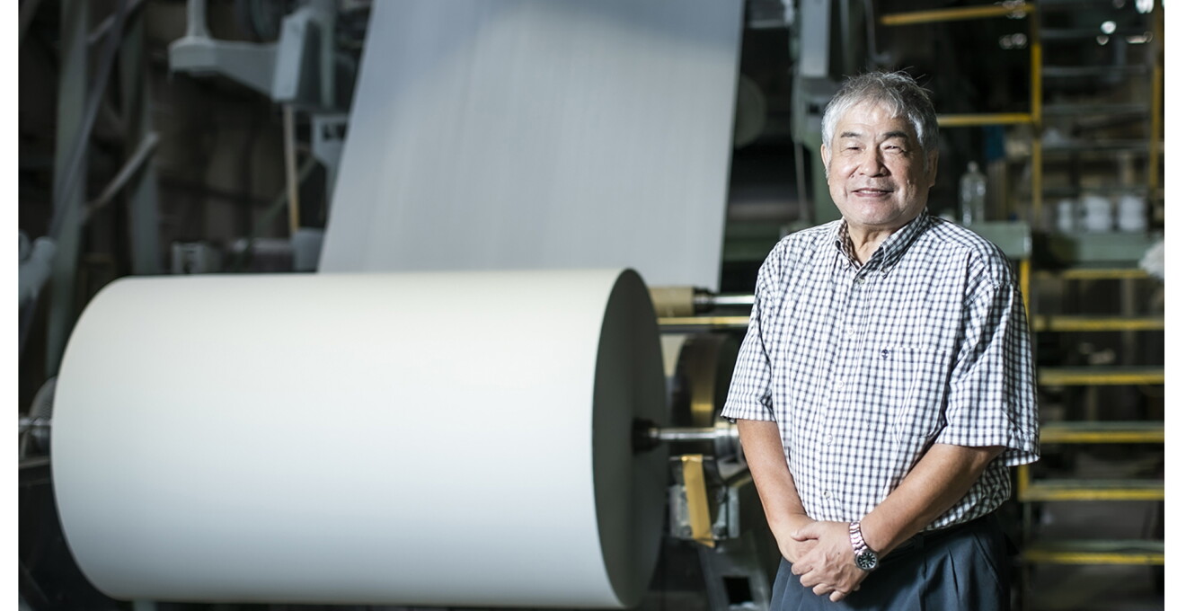 工場で製造中の大きなロール紙の横で、笑顔の太田さん