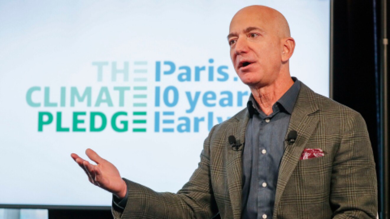De fondo el cartel the The Climate Pledge, Paris 10 years early. Jeff Bezos en primer plano con una mano levantada. Una amerciana verde y gris a cuadros y una camisa azul. En la americana lleva un pañuelo rojo.