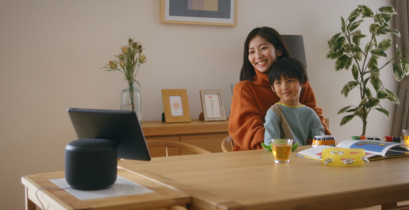 リビングのダイニングテーブルで、膝の上に子供をのせて座る女性。2人ともテーブル端のデバイスを見ている。机の上に開いた絵本やコップ。