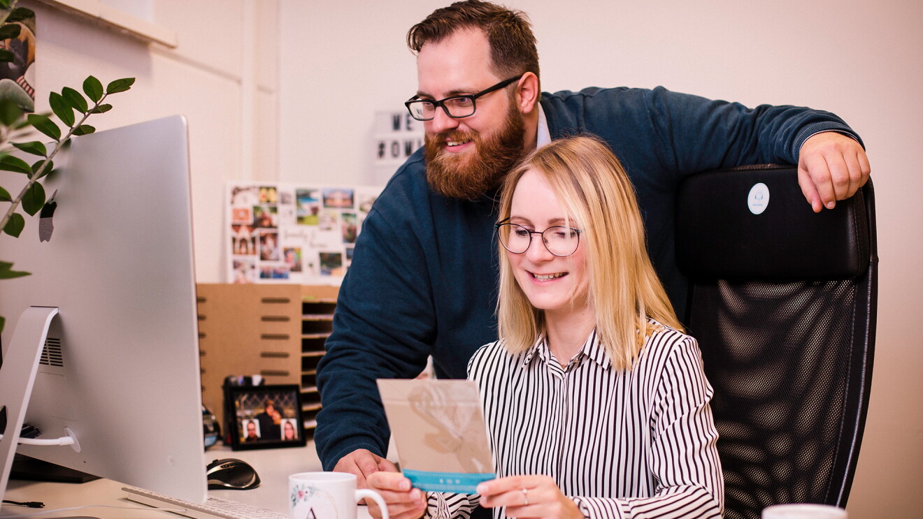 Eine Frau und ein Mann sitzen und stehen vor einem Computerbildschirm. Sie lächeln beide und schauen in den Bildschirm. Die blonde Frau hält eine Karte in den Händen, von der nur die Rückseite zu sehen ist. Sie befinden sich in einem Büro..