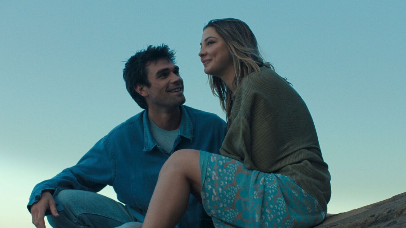 A scene from The Map That Leads to You with a couple sitting closely on rocky outcrop against blue sky