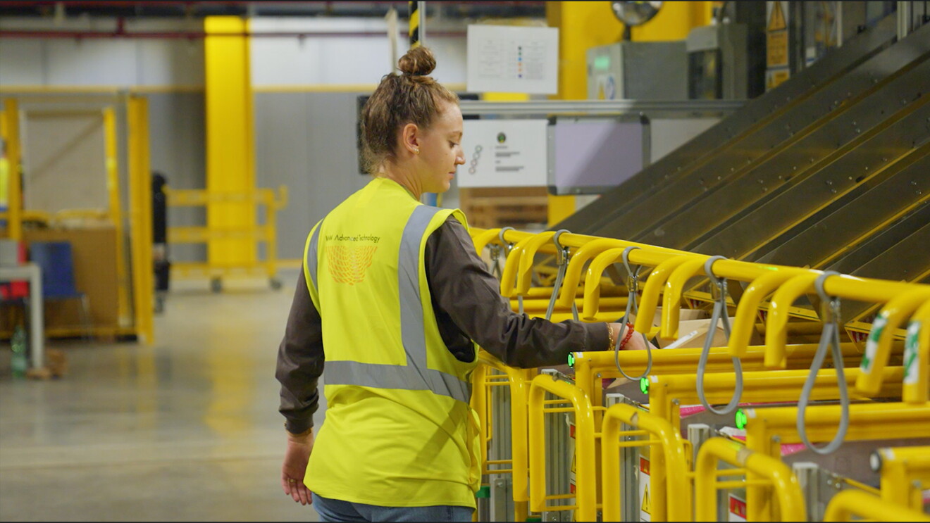Dentro de un centro logístico se encuentra una trabajadora cogiendo un sobre de la línea de envío. Fondo desenfocado con una estructura de color amarilla. La protagonista, con el pelo ondulado, recogido con un moño muy alto. Lleva un chaleco de color amarillo y una camisa de color verde oscuro. 