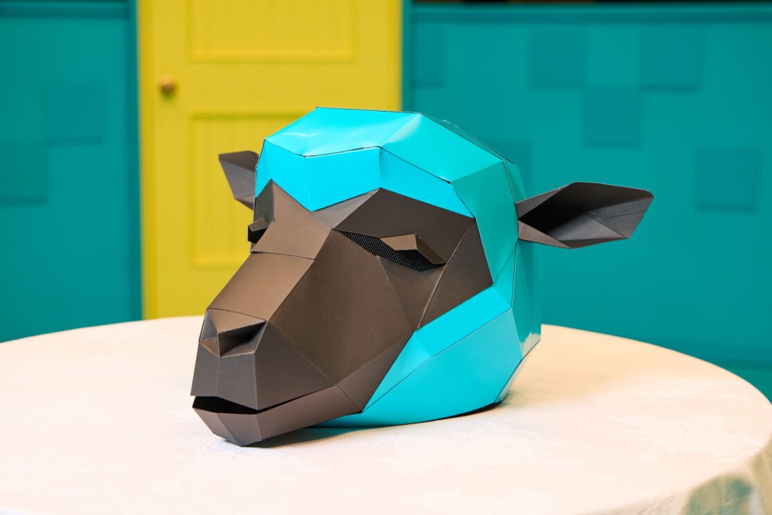 水色の壁と黄色いドアの手前に白いテーブルクロスを掛けた丸テーブルがあり、その上には、水色と黒の紙で作られた羊の仮面が置かれている。