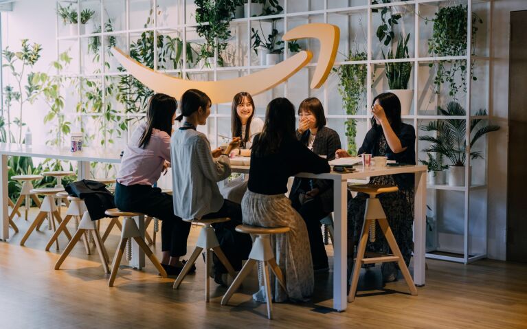 白い正方形で仕切られた棚の中に植物があり、その手前に大きな黄色いAmazonのスマイルロゴのレリーフがかけられている。その前に置かれたテーブルで6人の女性たちが話をしている