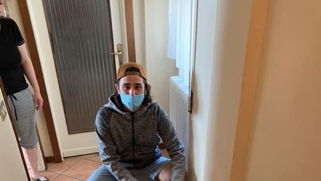 Un ragazzo sistema alcune scatole in un corridoio. Indossa una mascherina sul viso e un cappello in testa. E' inginocchiato per terra davanti alle scatole. 