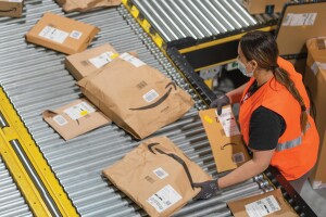 A woman wearing a safety vest stands to the right of a conveyor belt. She is handling flat packages, moving them along the belt.