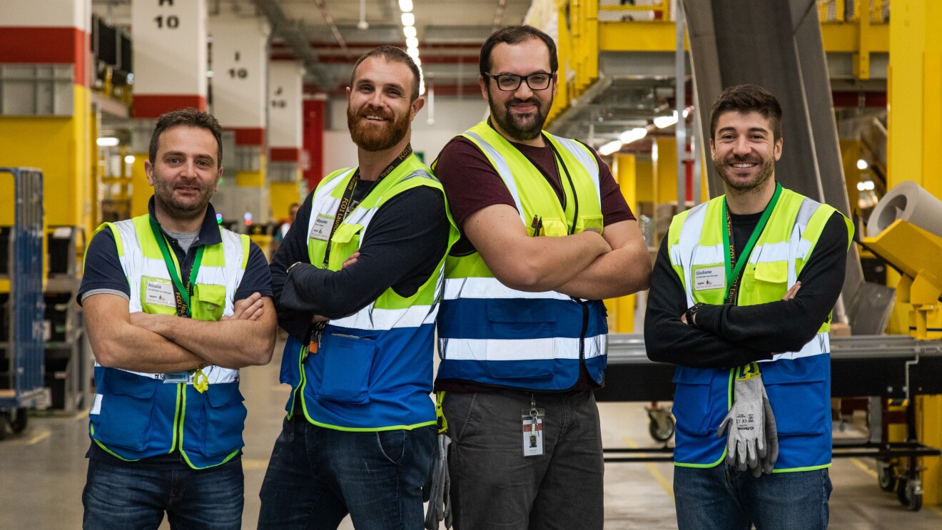 Quattro uomini con gilet ad alta visibilità e le braccia incrociate, guardano in camera e sorridono. Sono nel centro di distribuzione Amazon di Passo Corese.