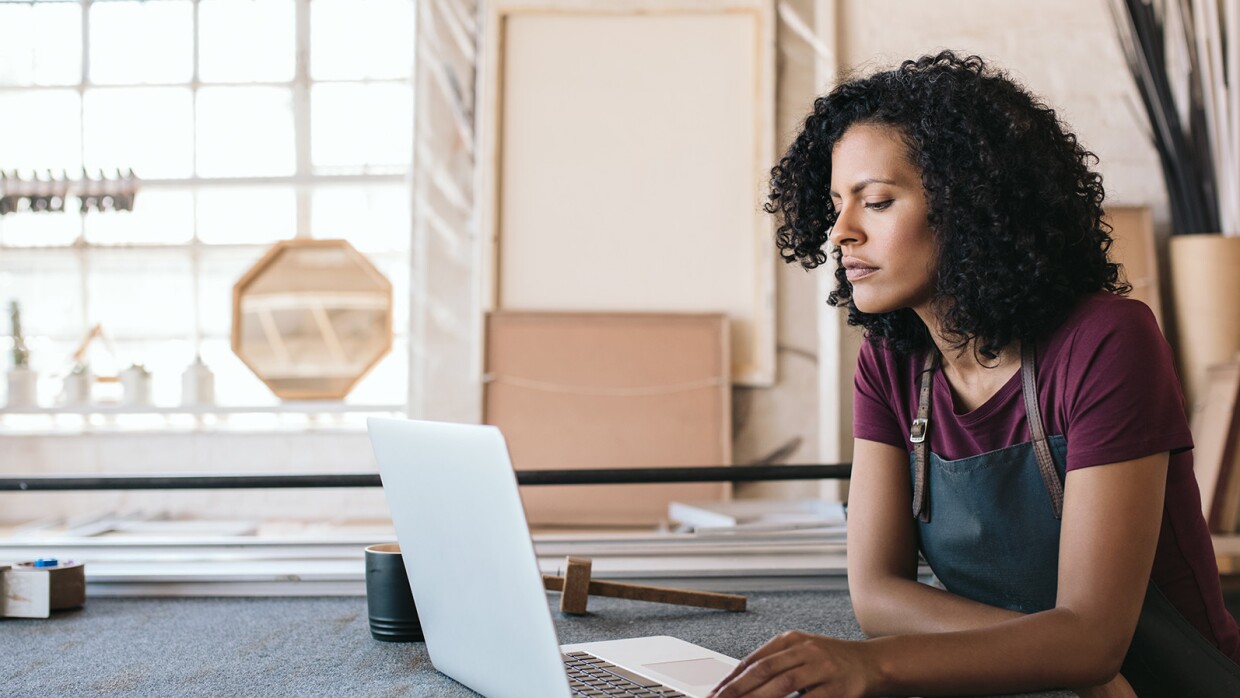 Un'imprenditrice di fronte al suo laptop in una bottega sta guardano lo schermo. Indossa un grembiule da lavoro, ha i capelli ricci.