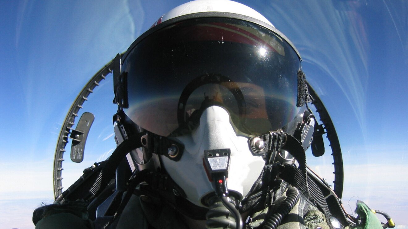 An image of a pilot flying a jet with a big window above them with a view of the blue sky. The pilot's face is covered with a large helmet, large black googles, and a breathing mask.