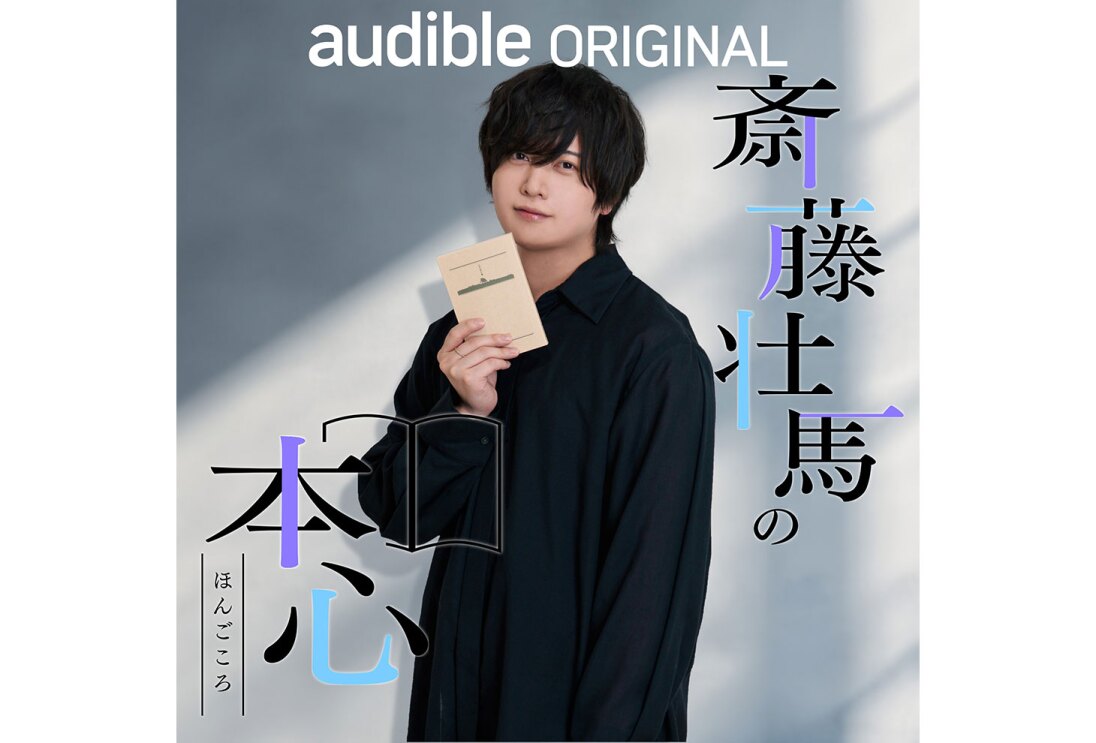 光が斜め右から差すグレーの壁を背景に立つ男性のポートレート。黒い服で、手に持った本を顔の近くに掲げている。男性の右側に「斉藤壮馬の」、左側に「本心」。上側に横書きで「audible ORIGINAL」。