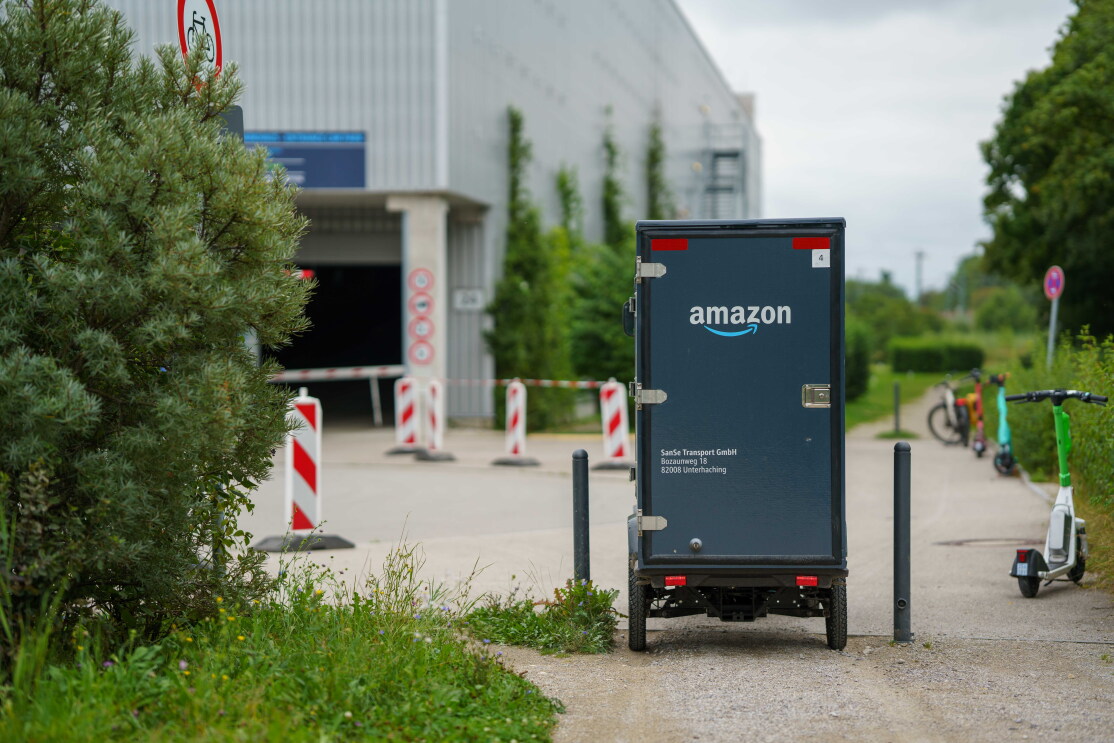 Ein blau-grauen E-Lastenrad fährt durch zwei Poller, im Hintergrund ist das Amazon-Verteilzentrum zu sehen.