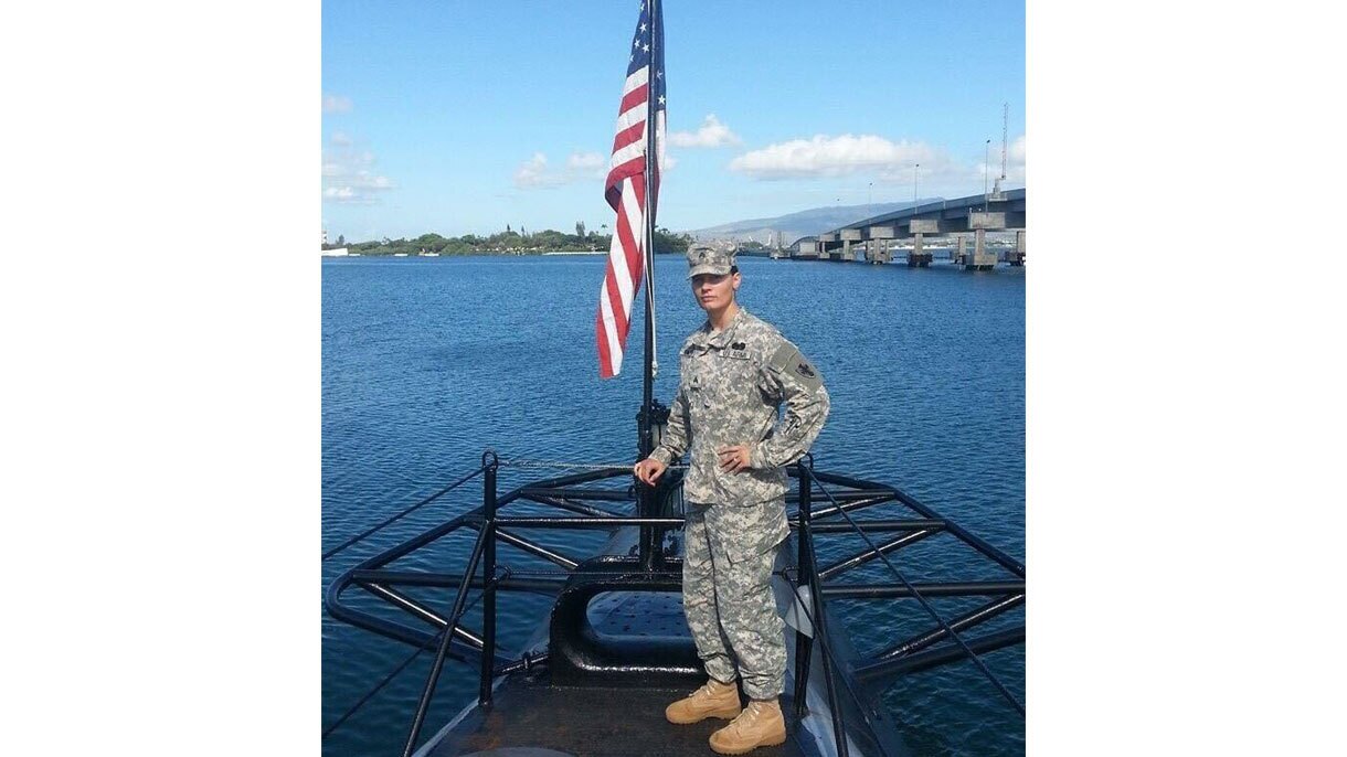 An image of Christina Carter in her military uniform standing for a photo on the front of a boat with the American flag in the background.