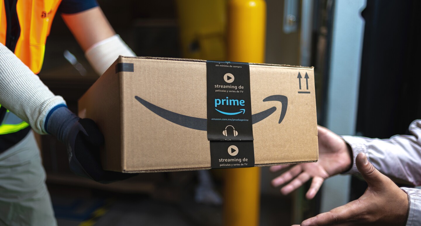 Primer plano de una caja de cartón de Amazon con las manos de un repartidor vestido con guantes negros y blanco u un chaleco de color naraja. Dos mano reciben la caja.