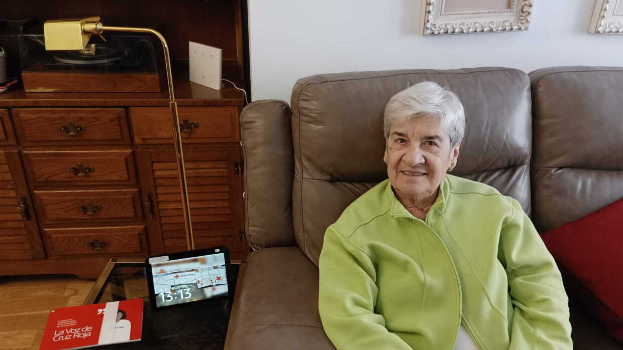 Mujer mayor sentada en el sofá con una mesita con Alexa y un documento de Cruz Roja.