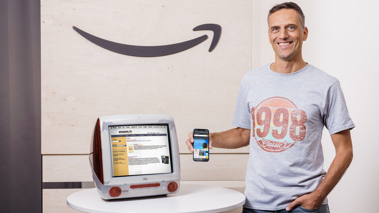 Amazon Deutschland gibt es seit 25 Jahren. Rocco Bräuniger hält ein Smartphone mit der Amazon App vor einen PC mit der Amazon Website.