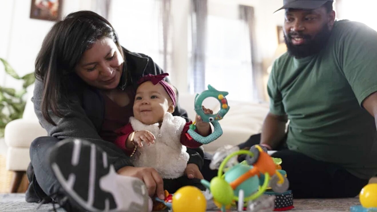 A woman sits with a baby surrounded by toys, while a man looks on.
