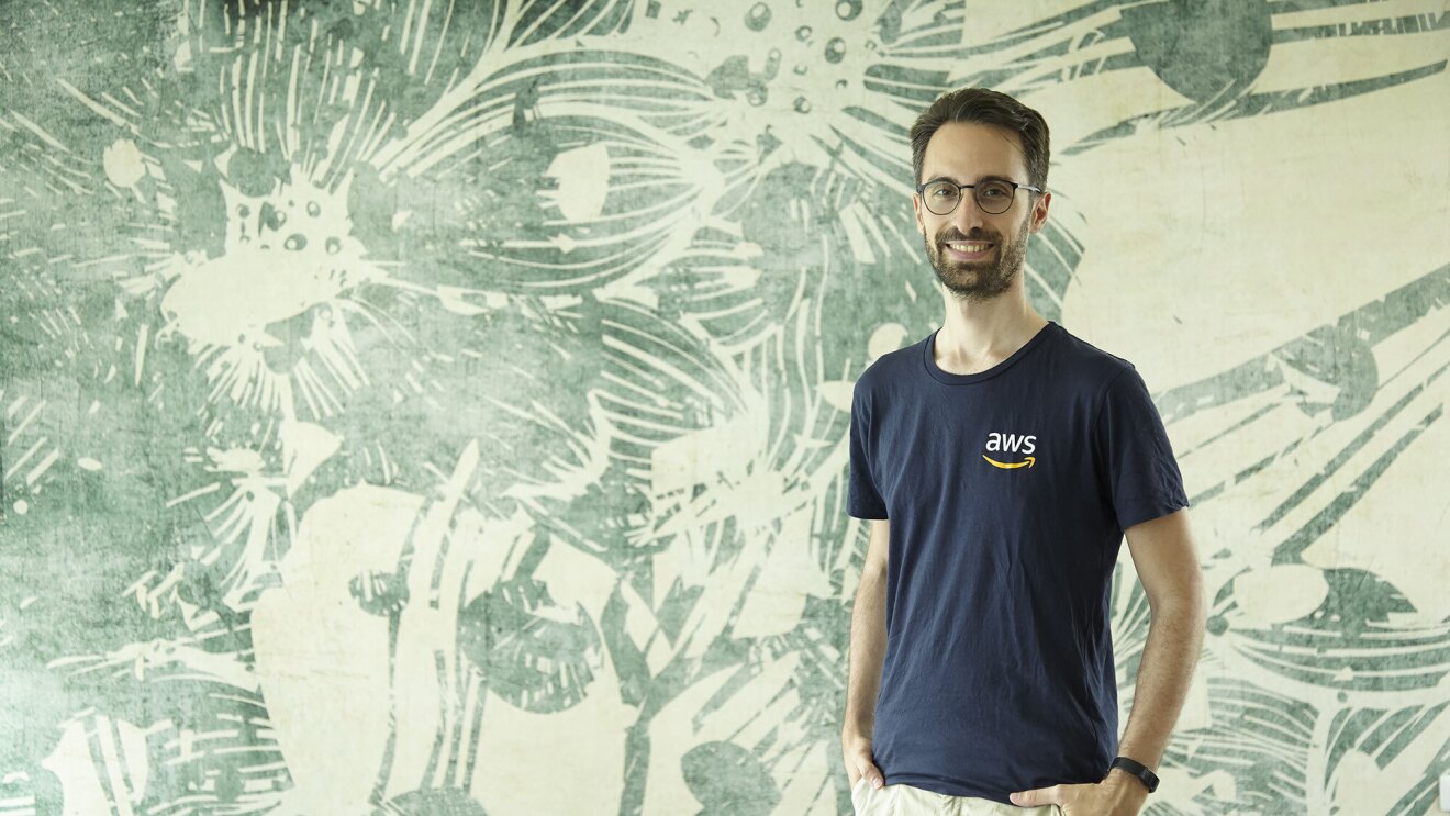 Mezzo busto di Alex Casalboni. Il ragazzo indossa una t-shirt blu con il logo AWS a destra, e un paio di occhiali. Sorride verso la camera. Sullo sfondo, una carta da parati beige e verde.