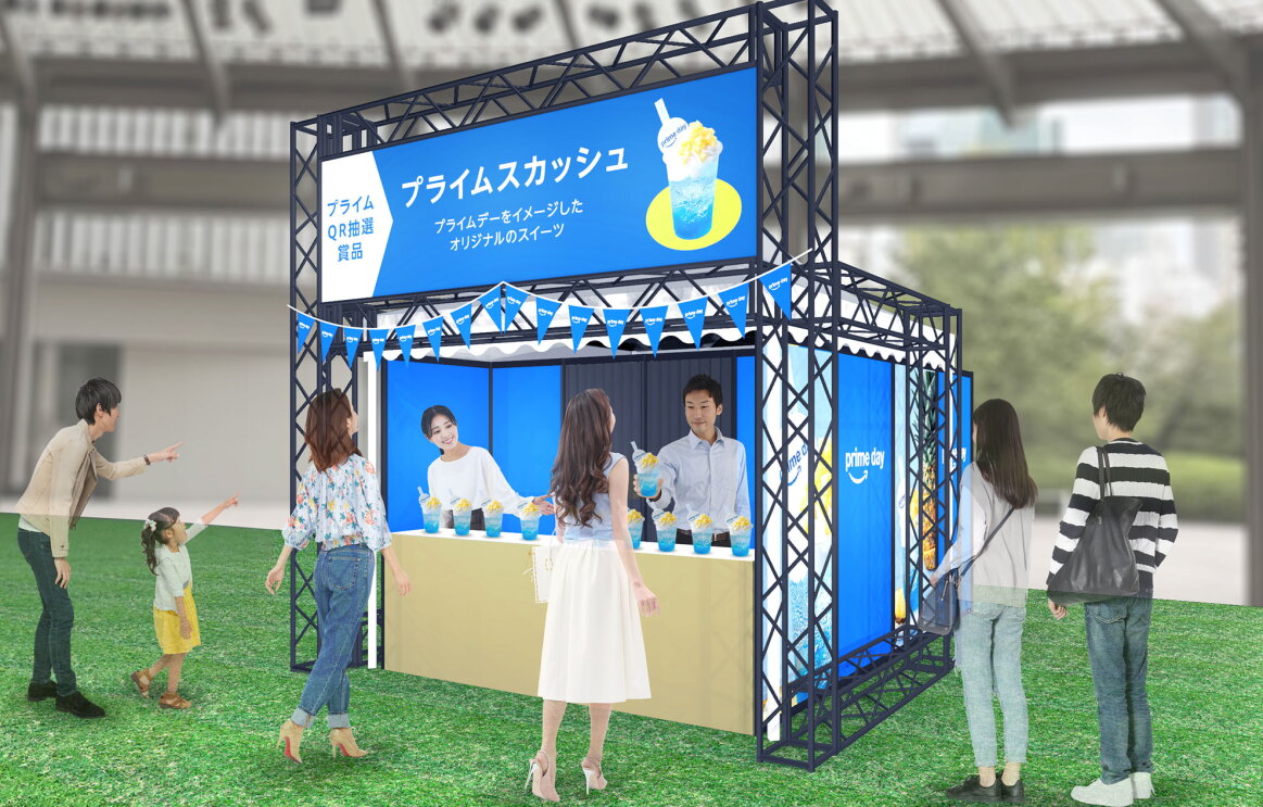 芝生の上に、青を基調とした販売ブースが設置されている。青い看板には白字で「プライムスカッシュ」と書かれ、飲み物のイラストが表示されている。ブースは青い三角旗で装飾されている。ブースを指さす親子や看板を見上げる人物などがいる。