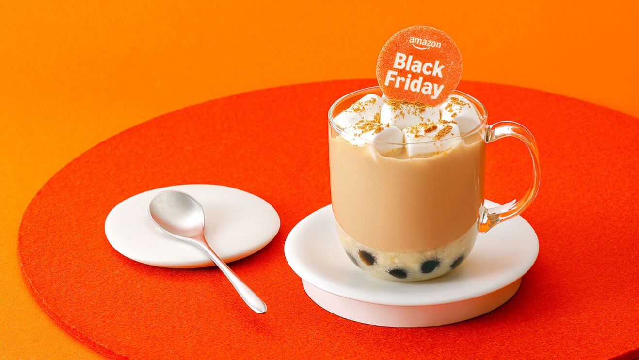 オレンジのテーブル、濃いオレンジの丸いマットに、ガラスカップでタピオカミルクティーを提供。下に白い受け皿、上部にマシュマロとオレンジの丸いプレートの装飾。左側に白い小皿と銀のスプーン。