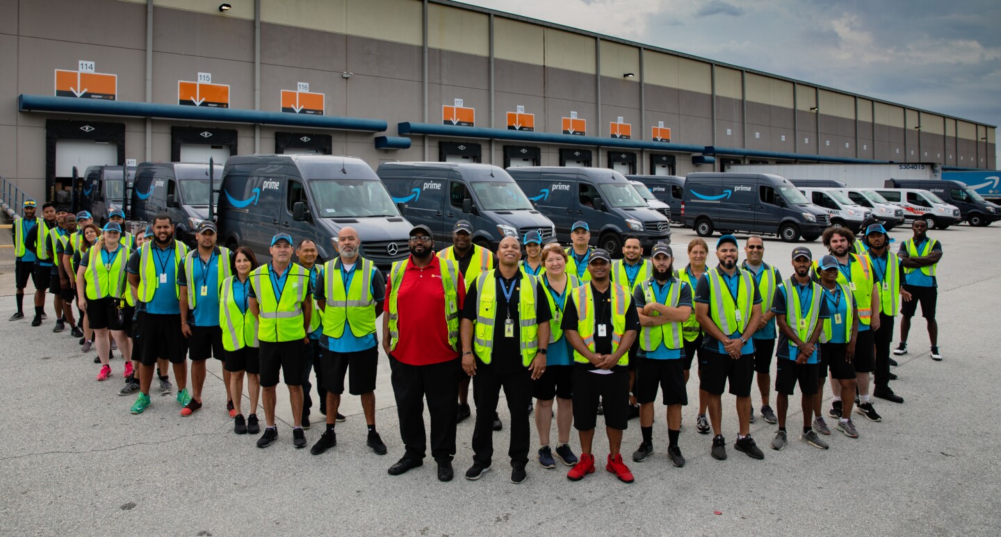 A group of employees stand in front of a fleet of Amazon Prime-branded cargo vans.