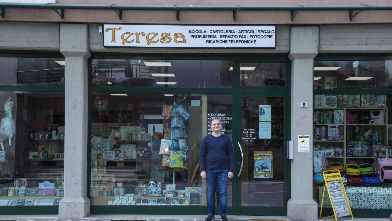 Andrea Locatelli posa di fronte all'ingresso del negozio di Teresa. L'insegna dice "Teresa - edicola, cartoleria, articoli regalo, profumeria, servizio fax, fotocopie, ricariche telefoniche."