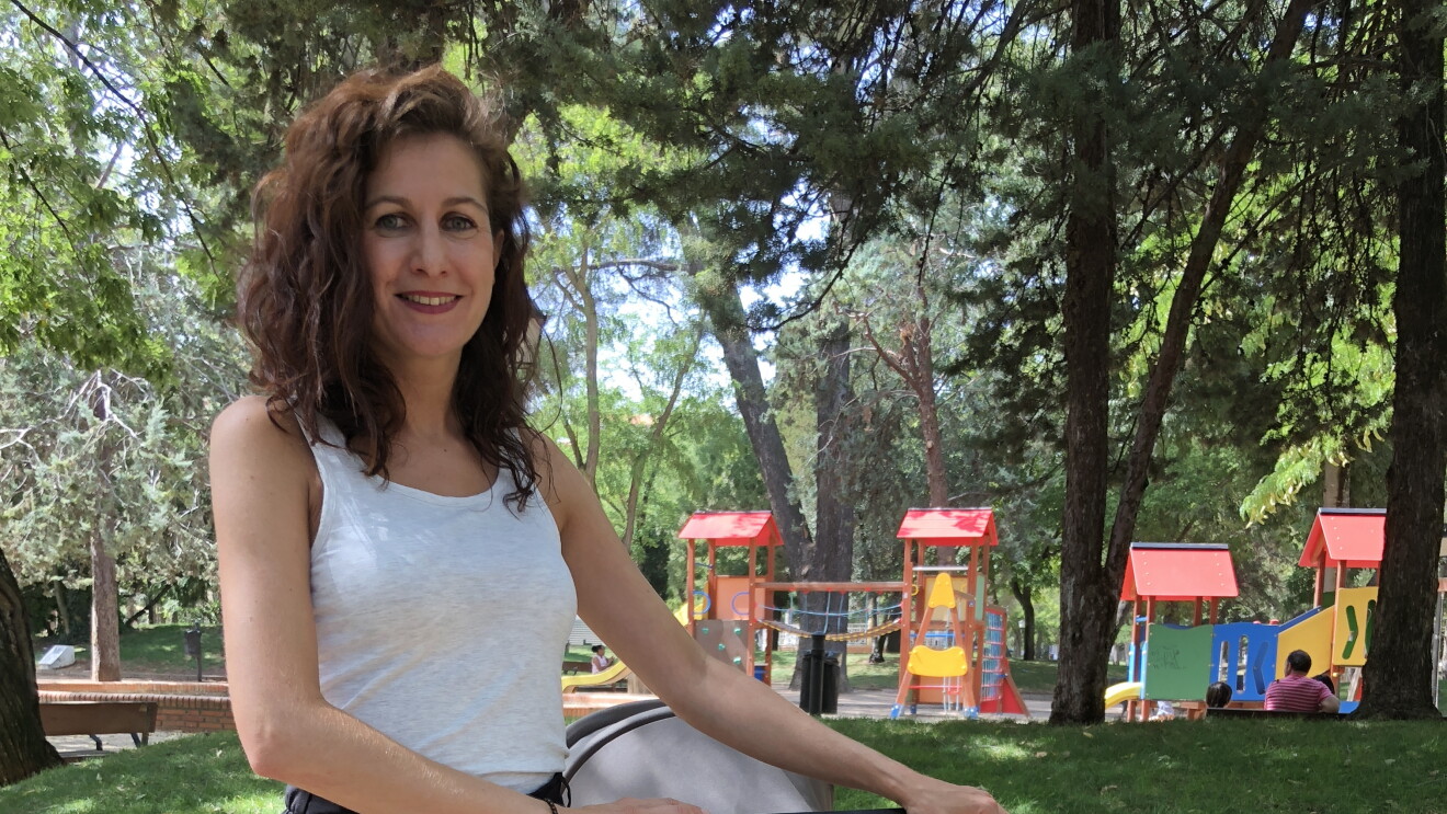 Irene posa de pie sujetando un carro de bebé que tiene su bolso, Andone Dynamic Walks. Mira a cámara sonriendo y va vestida con una camiseta blanca sin mangas y una falda larga. Está en un parque y en el fondo se puede ver una zona de juegos infantil.