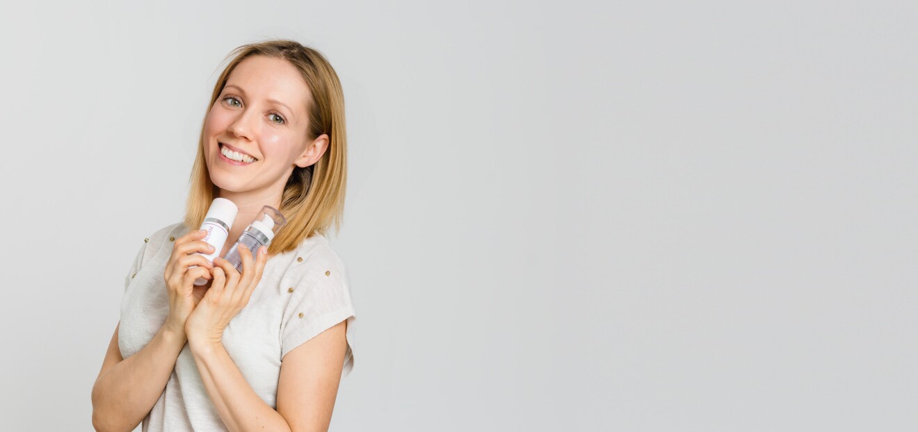 Eine Frau mit blonden Haaren und einem weißen T-shirt lächelt in die Kamera. In ihren Händen hält sie zwei Kosmetikflaschen.