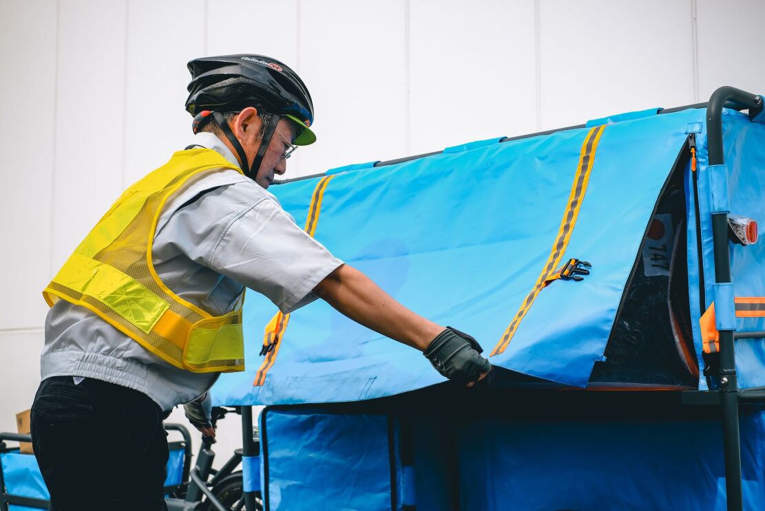 ヘルメットを被っている男性がリアカーの青いカバーを閉じている。その中には大きな荷物が見える