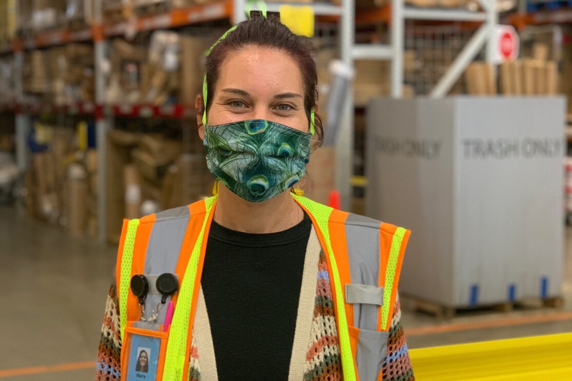 A woman wearing  a safety vest and a mask that covers her mouth and nose stands in a space with tall shelves in the background.