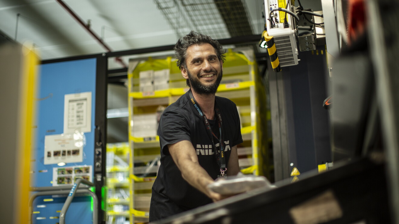 Elia works in a FC. He has a black tshirt, a big smile and he is picking an item. ELIA VILLARDITA - Operatore di magazzino :"Sicurezza sul lavoro significa creare un ambiente lavorativo dove non ci sono condizioni di pericolo. Avere sicurezza sul lavoro porta tranquillità, è bello sapere che i rischi sono quasi nulli, questo ci permette di lavorare meglio e quindi in modo più produttivo".