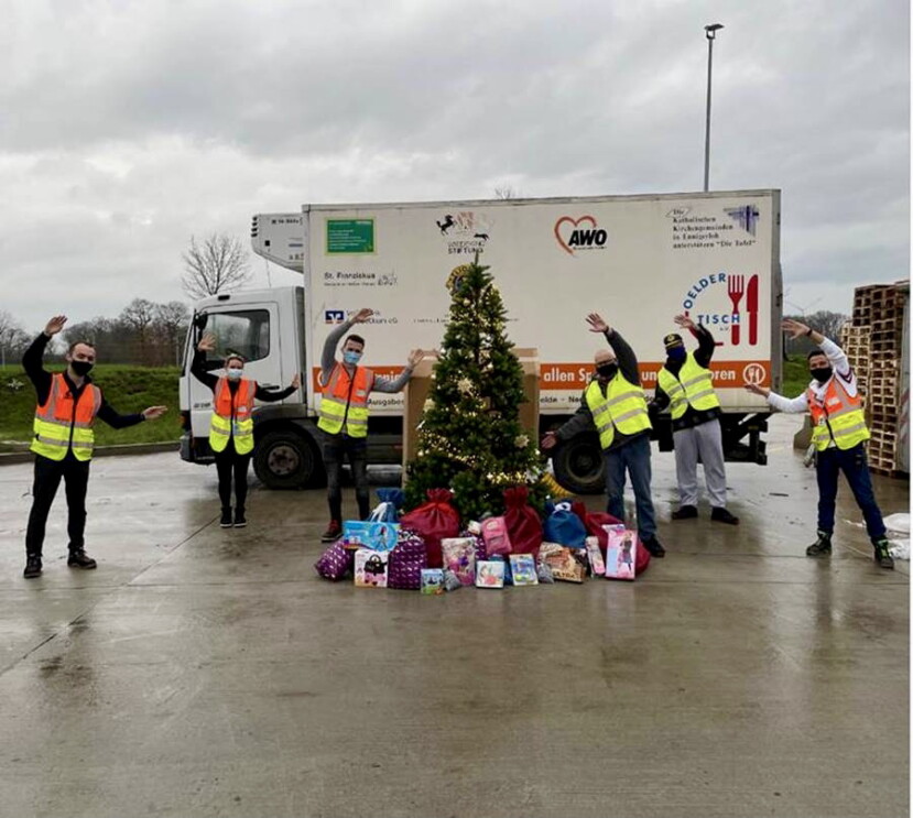 Gruppenbild mit Mitarbeiter von Amazon und der Tafel vor einem Tafel-Lkw. In der Bildmitte ein Weihnachtsbaum, unter dem Geschenke liegen.