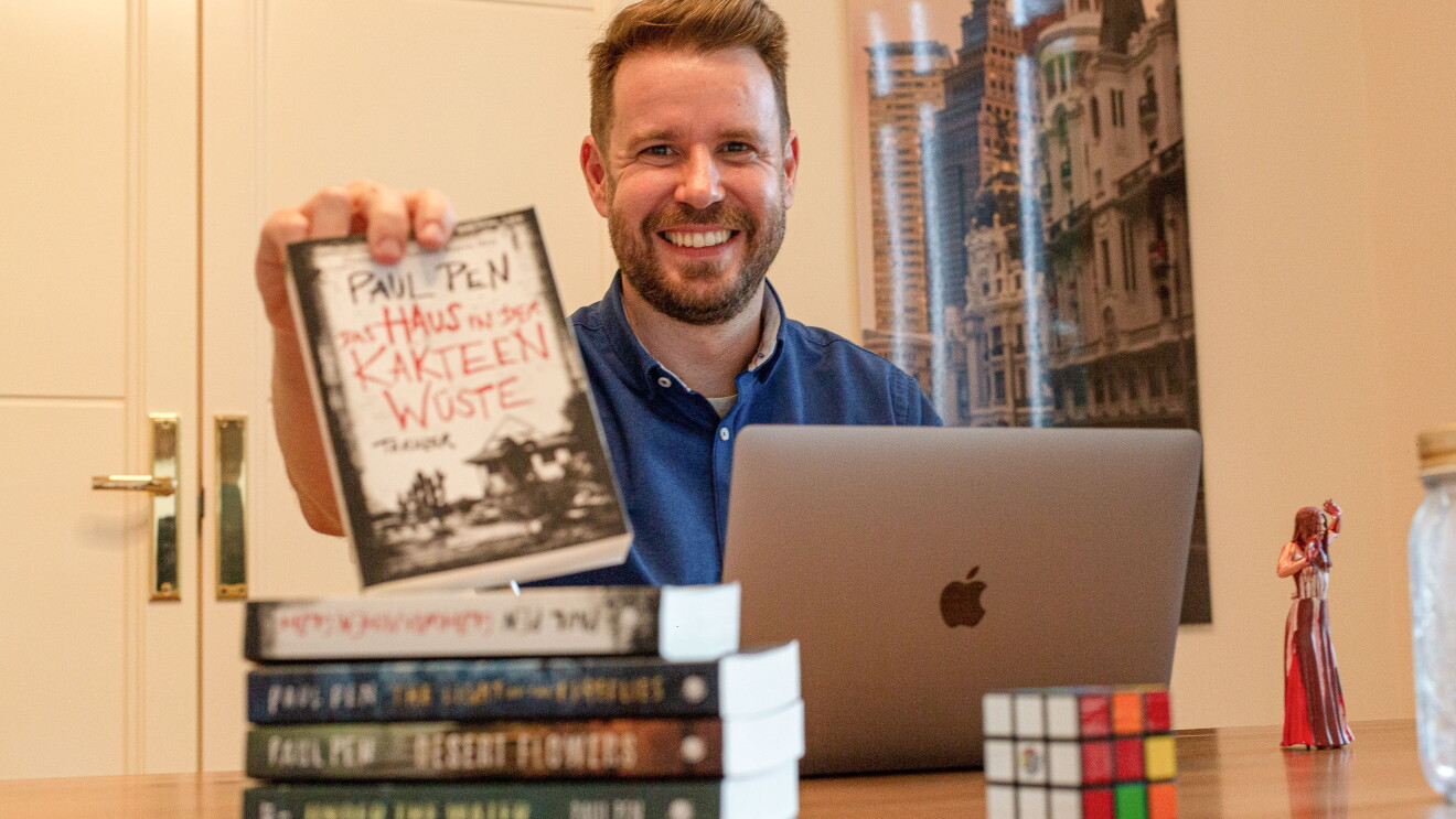 Paul Pen mostrando la traducción al alemán de su novela La casa entre los cactus. En su habitación de trabajo Paul sujeta su novela traducida al alemán con su mano derecha. Delante tiene el ordenador, sus otras novelas traducidas. De fondo hay una puerta y un poster colgado en la pared. Paul está mirando sonriendo a cámara con una camisa azul. 