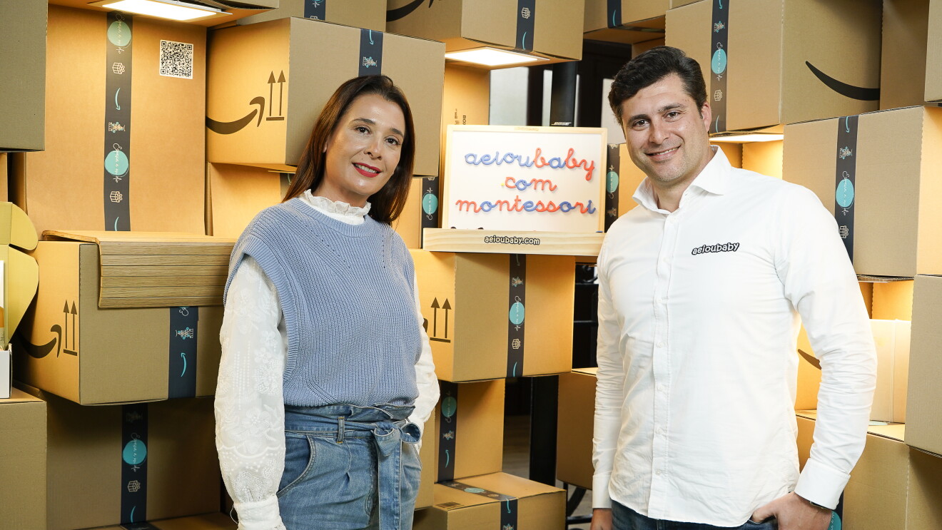 En un escenario formado por cajas de Amazon de cartón. Marga López y Lucas Díez son los fundadores de AEIOU Baby, una pyme de Talavera de la Reina (Toledo) que, desde que nació en 2016, vende productos exclusivamente en Amazon. Marga va con unos jeans azules de tiro alto con cinturón de tela, atado en un lazo en la cintura. Debajo una camisa y un jersey sin mangas azul. Lucas lleva una camisa blanca con el nombre de la empresa AEIOU Baby. Lleva el pelo corto, jeans i las mano en apoyados en los bolsillos. Entre ellos y apoyados en una caja de Amazon, la pizarra de madera con letras azules y rojas y el texto aeioubaby.com Montessori. 