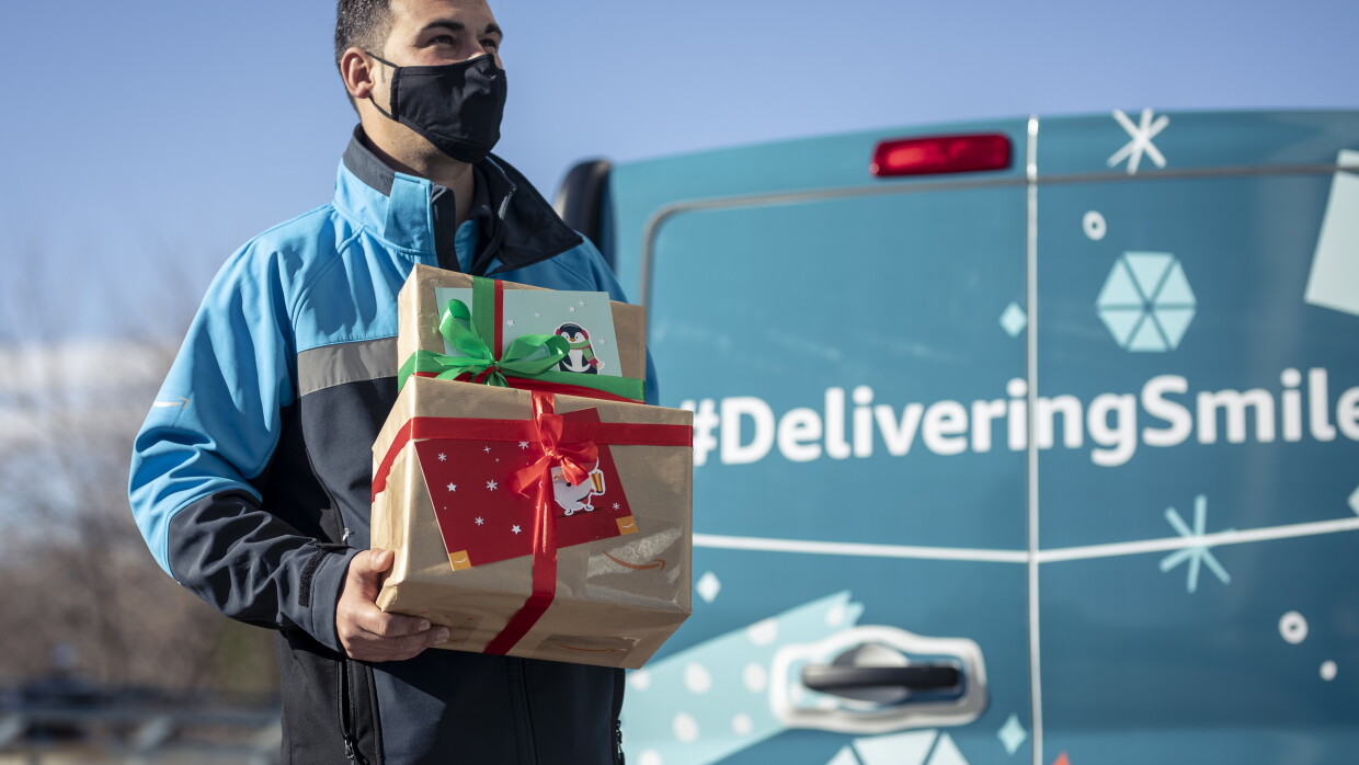 Donación al Centro de día de personas sin hogar Casablanca de Alcobendas.  A la parte derecha de la foto y en segunda plano, una furgoneta de Amazon de color azul-verde. Se ve solo la parte trasera y tiene la etiqueta impresa de DeliveringSmiles. En primer plano un repartidor de Amazon con pelo negro corto, mascarilla negra y un anorac de color azul claro, azul marino y gris. Carga dos cajas de Amazon envueltas en papel marrón con un lazo una rojo y la otra verde y dos tarjetas regalo. Mira al horizonte y se le imagina una sonrisa en el rostro. 
