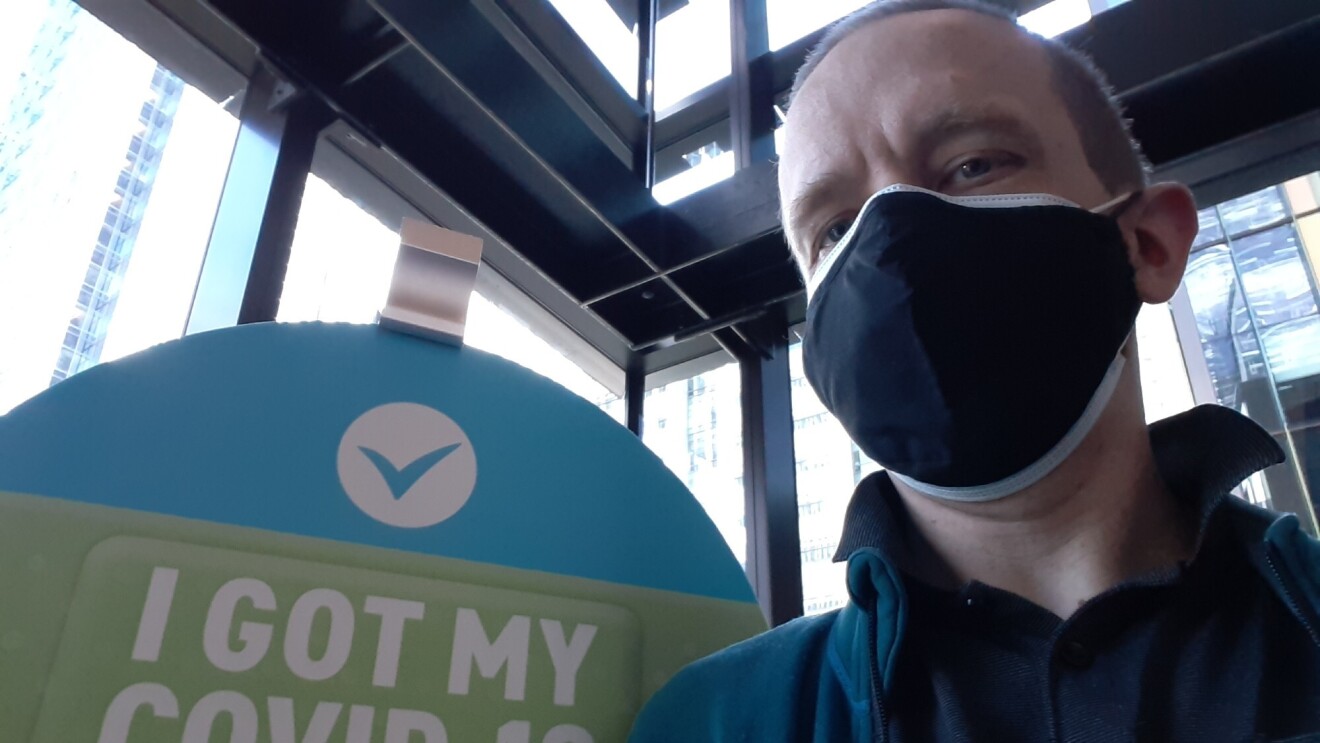 一个男人戴着面具,一件衬衫坐在前面的说明说“我得到了我的COVID-19疫苗。”