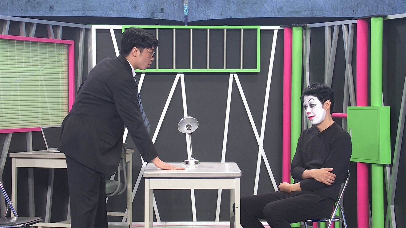 黒い壁、窓枠やパイプが黄緑とピンクに塗られた取調室のセットで、2人の男性が机を挟んで対峙。左側は黒いスーツで立って机に手を置き、右側は黒の上下にピエロのメイクアップで椅子に腰かけている。