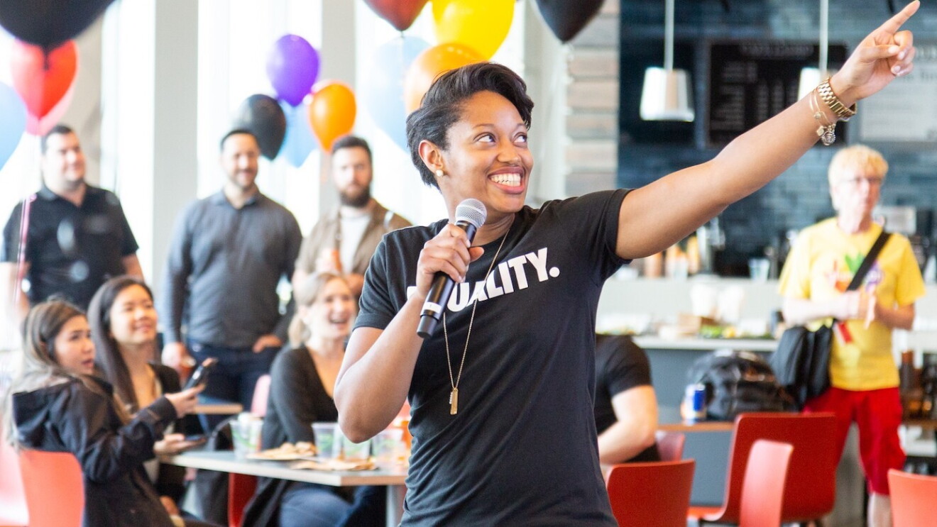 A Black woman wearing a shirt that says "Equality" speaks into a microphone while gesturing off camera. She is speaking to a brightly decorated room full of people.